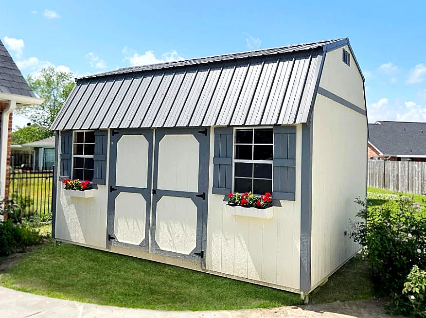 Cream-painted Derksen Portable Building lofted barn with grey metal gambrel roof, grey shutters, white window flower boxes with red flowers, displayed on a Larkin's Portable Buildings Nebraska lot.